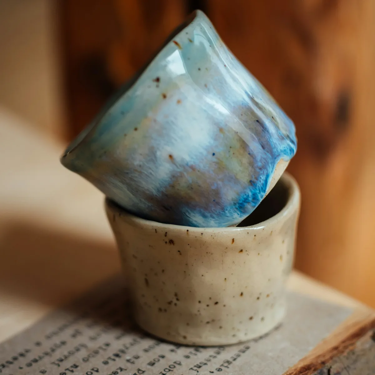 Two handmade ceramic cups, a blue glazed cup stacked on a speckled one, photographed on an open book against a wooden shelf